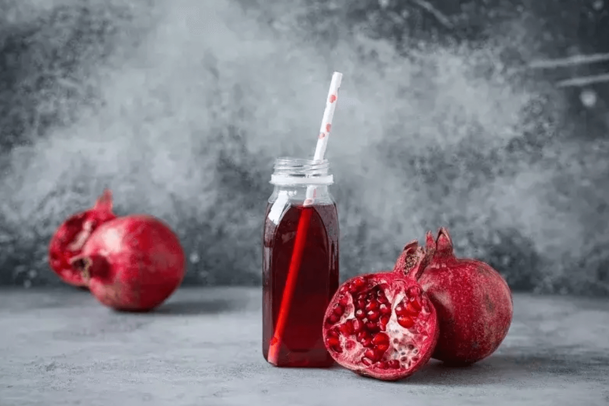 Pohár granátovej šťavy so slamkou, obklopený celým a rozpoleným granátovým jablkom na sivom pozadí – symbol prírodného antiparazitiká.
