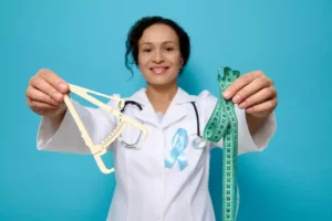 Lekár alebo odborník v bielom plášti drží v jednej ruke kaliper na meranie podkožného tuku a v druhej ruke krajčírsky meter – symbol vyšetrenia obezity a rizík metabolického syndrómu.