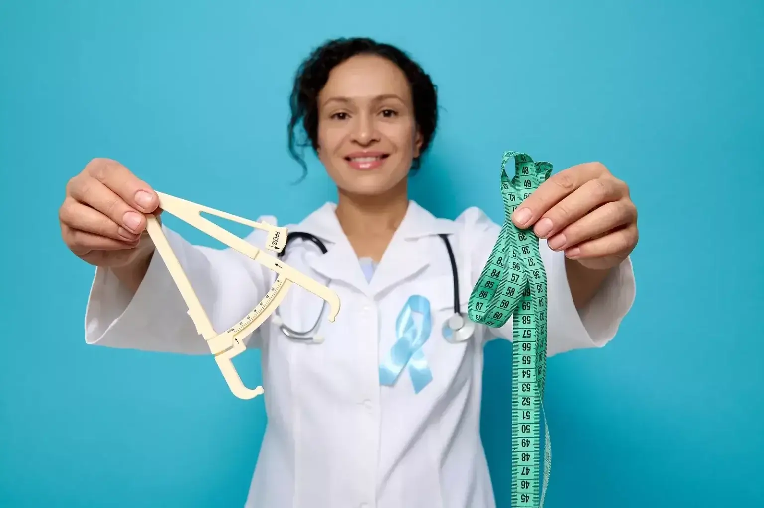 Lekár alebo odborník v bielom plášti drží v jednej ruke kaliper na meranie podkožného tuku a v druhej ruke krajčírsky meter – symbol vyšetrenia obezity a rizík metabolického syndrómu.