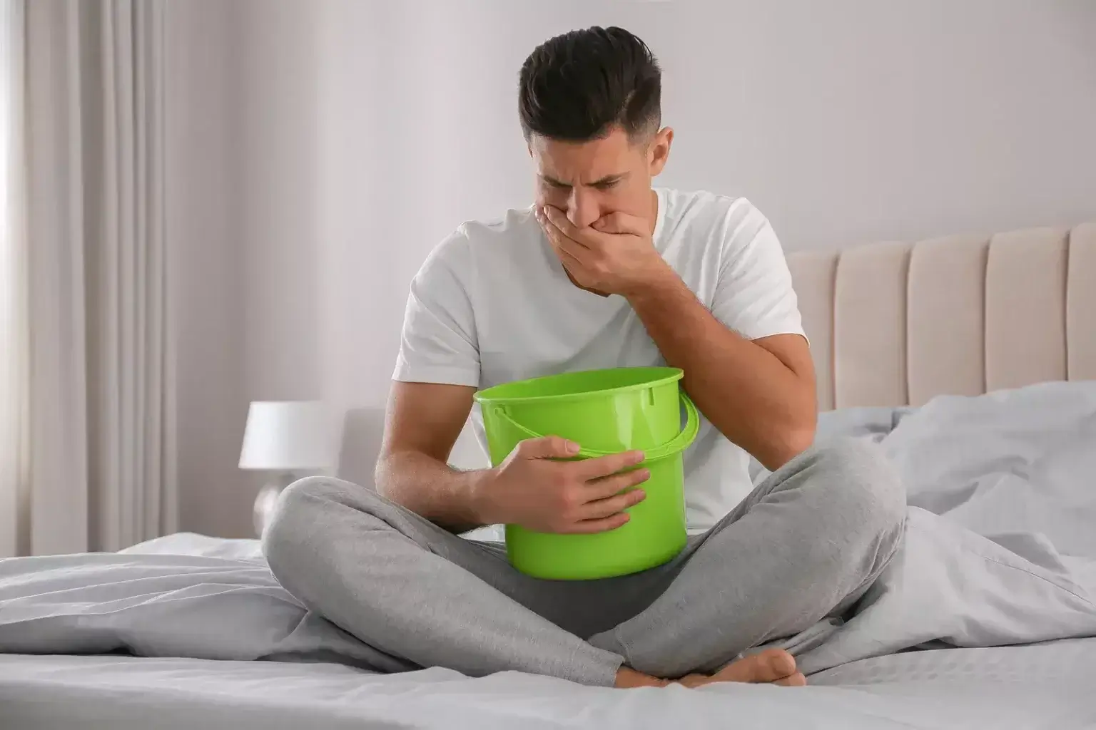 Muž v bielom tričku sedí na posteli so zeleným vedrom v rukách, vyjadruje nevoľnosť a nutkanie na zvracanie – príznaky tráviacich ťažkostí.