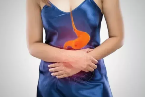 Žena v modrých šatách si drží brucho, zvýraznený pažerák a žalúdok s naznačeným zápalom – ilustrácia symptómov zápalu pažeráka (ezofagitídy).