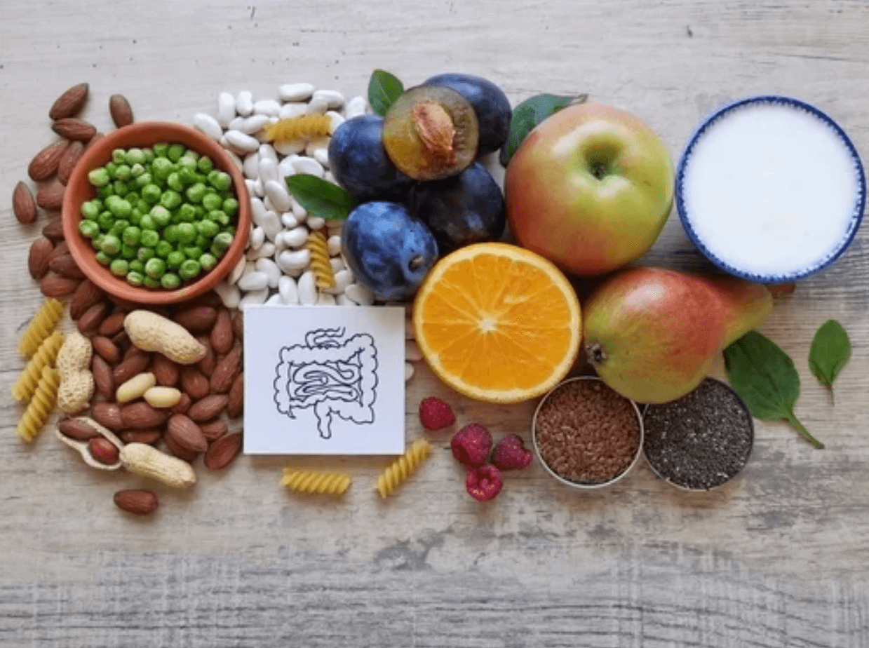 Rôzne potraviny bohaté na beta-glukán, ako zelenina, ovocie, orechy, strukoviny, semienka a jogurt, zoradené na stole spolu s ilustráciou čreva – symbolizujúc účinky beta-glukánu na zdravie tráviaceho systému a podporu imunity.
