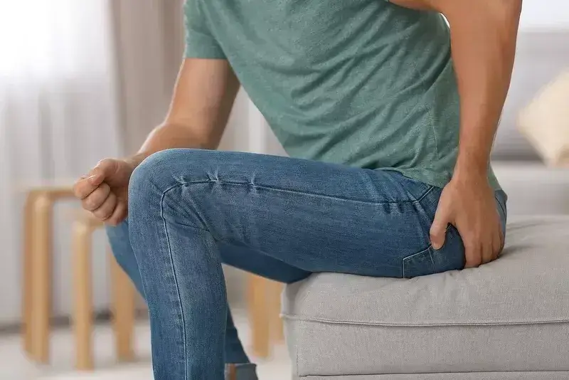 Muž v rifliach si drží ruku na zadnej časti tela v oblasti konečníka, v druhej ruke drží toaletný papier – vizuálne vyjadrenie bolesti alebo diskomfortu typického pre hemoroidy.