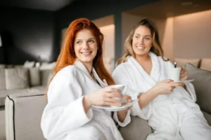 Dve ženy v bielych županoch oddychujú na pohovke a držia šálky s čajom – atmosféra domácej relaxácie a pohodlia počas detoxikácie.