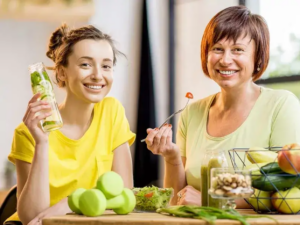 Dve ženy sediace za stolom s miskou a tanierom plným čerstvého ovocia a zeleniny, jedna drží pohár so zeleným smoothie, druhá naberá šalát vidličkou – symbol zdravého životného štýlu a výživy pri detoxikácii organizmu.