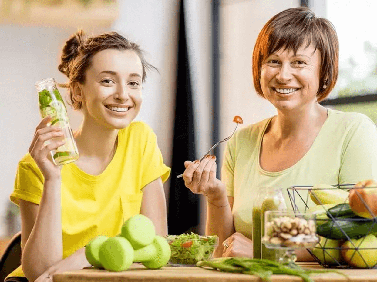 Dve ženy sediace za stolom s miskou a tanierom plným čerstvého ovocia a zeleniny, jedna drží pohár so zeleným smoothie, druhá naberá šalát vidličkou – symbol zdravého životného štýlu a výživy pri detoxikácii organizmu.