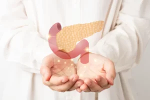 Osoba v bielom oblečení jemne drží v dlaniach digitálnu ilustráciu pankreasu so zvýraznenými bodmi – symbolizuje dôležitosť zdravia slinivky, jej anatomickú polohu a prevenciu ochorení.