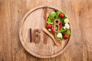 Hodiny vyskladané z príboru a čerstvej zeleniny s číslom 16 a 8 znázorňujúce princíp prerušovaného pôstu – vizuálna metafora časovo obmedzeného stravovania pre podporu detoxikácie organizmu.