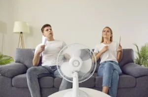Muž a žena relaxujú na pohovke, počas horúceho letného dňa sa ochladzujú ventilátorom a vejármi v interiéri, ilustrujúc dôležitosť oddychu a hydratácie počas horúčav.