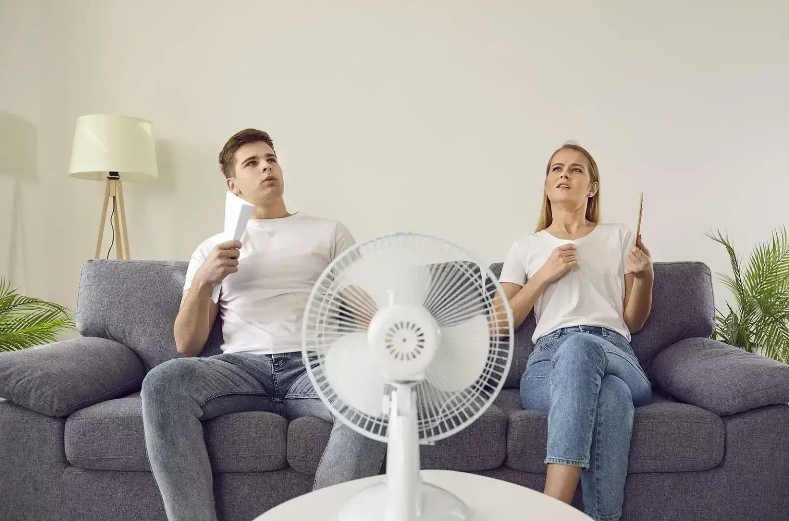 Muž a žena relaxujú na pohovke, počas horúceho letného dňa sa ochladzujú ventilátorom a vejármi v interiéri, ilustrujúc dôležitosť oddychu a hydratácie počas horúčav.